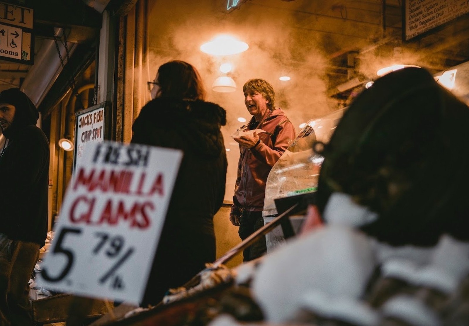 A middle aged man with his left hand raised to shoulder height is in focus in the background with an out of focus sign for manila clams in the foreground