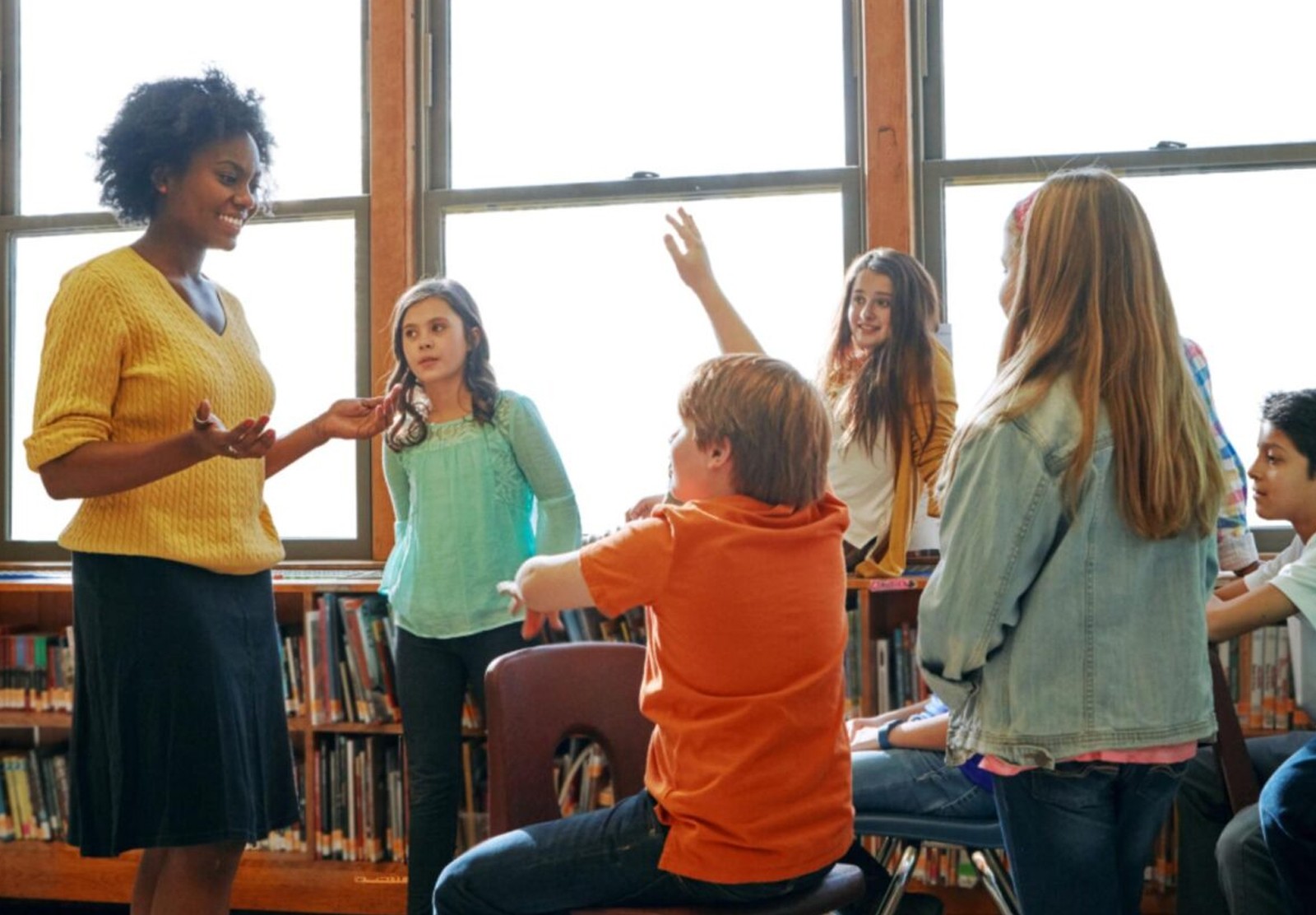 A woman in a yellow sweater is standing in front of a group of students, one of whom has their hand raised